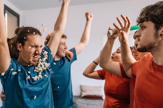 group of friends celebrating a soccer championship on TV. losing team throws popcorn at the winning team. leisure concept. happy and cheerful. natural light in the living room at home. trumpet