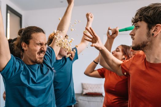 groupfriends celebrating a sports championship on TV. losing team throws popcorn at the winning team. e-sports. soccer. leisure concept. happy and cheerful. natural light in the living room at home. trumpet