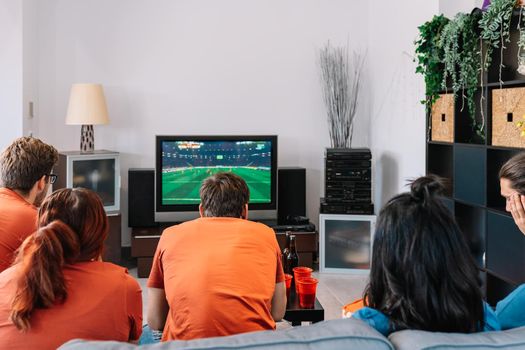 football friends celebrating a victory at home. young people watching sport on TV. leisure concept, three young adults in blue t-shirts. happy and cheerful. natural light in the living room at home.