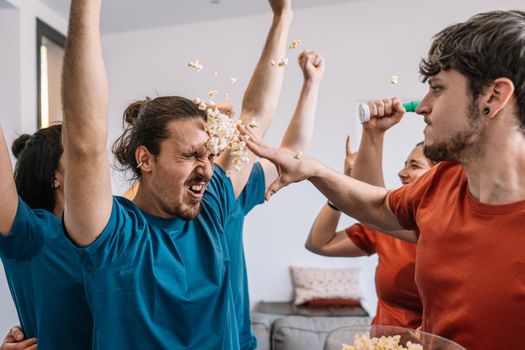 group of friends celebrating a e-sports championship on TV. losing team throws popcorn at the winning team. leisure concept. happy and cheerful. natural light in the living room at home. trumpet