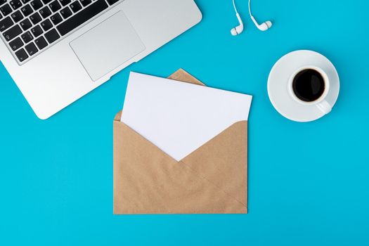 Open craft envelope with white blank paper sheet on bright blue background. Laptop keyboard, cup of dark espresso and earphones. Concept of receiving letter mail