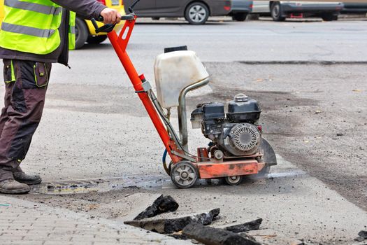 A road worker in reflective overalls uses a portable asphalt cutter to cut worn asphalt with a diamond blade to repair part of the roadway.