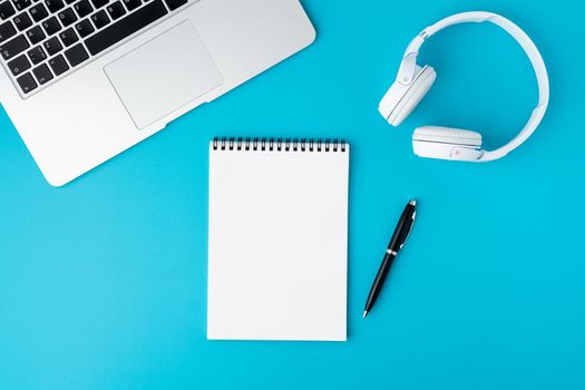 Blank empty spiral notebook on bright blue background. Laptop keyboard, headphones and automatic pen. Concept of modern workplace flat lay. Portable office equipment. Wireless technology, horizontal