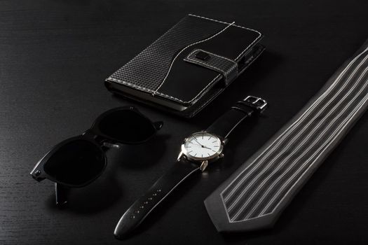 Watch with a leather strap, notebook in leather cover, sunglasses, silk tie on a black background