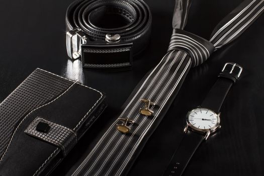 Notebook in leather cover, tie, cufflinks, leather belt with metal buckle, watch with a leather strap on a black background
