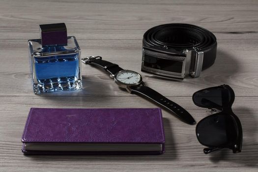 Man perfume, watch with a leather strap, leather belt with metal buckle, notebook in purple cover, black sunglasses on a gray wooden background