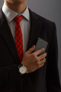 Young man in black suit keeping his hand with wristwatch on his chest and holding a phone