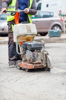 A road worker in reflective clothing cuts old asphalt on the road with a gasoline cutter against the background of a city street in blur. Copy space, vertical image.