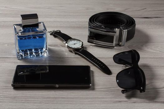 Man perfume, watch with a leather strap, leather belt with metal buckle, sell phone, black sunglasses on a gray wooden background