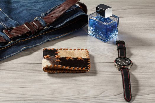 Man perfume, watch with a leather strap, jeans with leather belt and leather purse on a gray wooden background