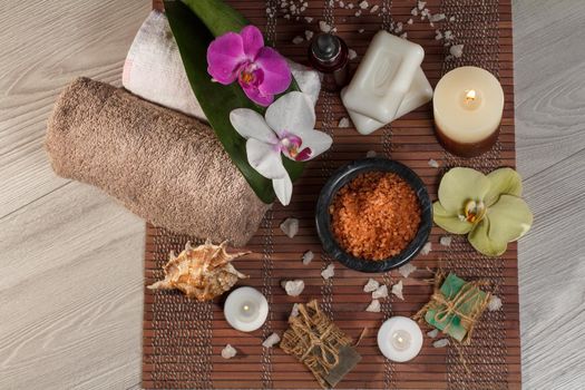 Spa and wellness setting with orchid flowers, bowl with sea salt, seashell, bottle with aromatic oil, soap, candles and towels on bamboo napkin