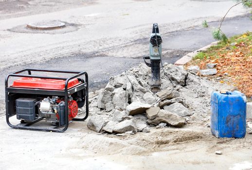 Electric jackhammer with a gas generator, a blue can of gasoline on the background of a workplace with a road section being repaired, copy space.