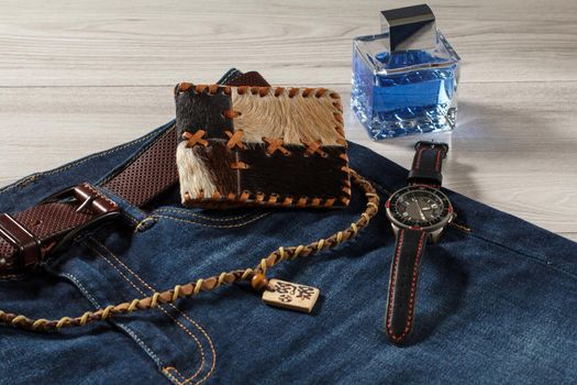 Man perfume, watch with a leather strap, jeans with leather belt, leather purse and amulet on a gray wooden background