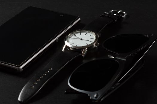 Watch with a leather strap, sell phone, black sunglasses on a black background