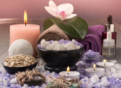 Spa treatment products with orchid flower, bowl with sea salt, bottles with aromatic oil, soap, scrub, candles and towels on wooden board