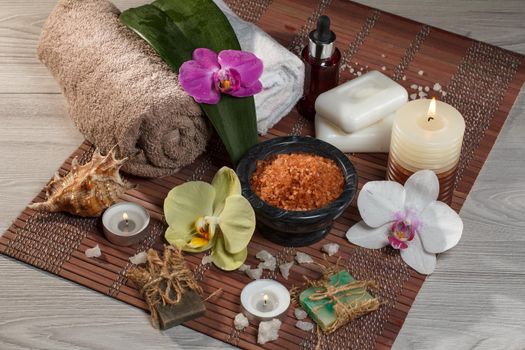 Spa and wellness setting with orchid flowers, bowl with sea salt, seashell, bottle with aromatic oil, soap, candles and towels on bamboo napkin