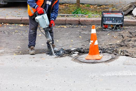 A road worker in reflective clothing smashes old asphalt with an electric jackhammer near a manhole.