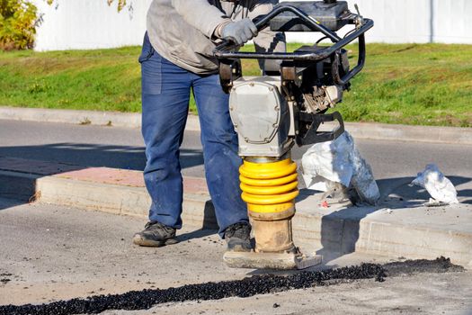 A road worker uses a petrol vibratory rammer to repair asphalt on the roadway on a bright sunny day.