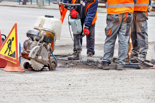 Three road service personnel in reflective clothing on a fenced-in section of road repair the roadway using a hand-held road tool and a jackhammer.