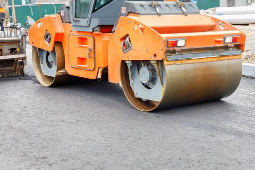 The metal cylinders of the large vibratory roller roll on the new road surface after the hot asphalt operator has poured the asphalt, providing a powerful compaction of the new road.