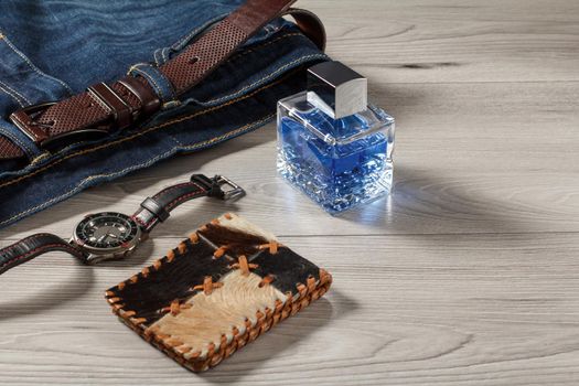 Man perfume, watch with a leather strap, blue jeans with leather belt and leather purse on a gray wooden background