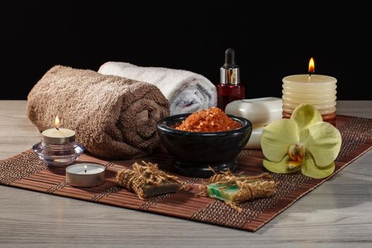 Spa and wellness setting with orchid flower, bowl with sea salt, bottle with aromatic oil, soap, candles and towels on bamboo napkin