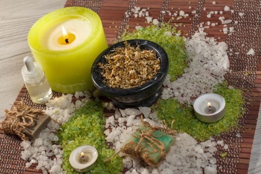 Spa accessories with soap, black bowl with dried chamomile flowers, bottles with aromatic oil, sea salt, candles on bamboo napkin