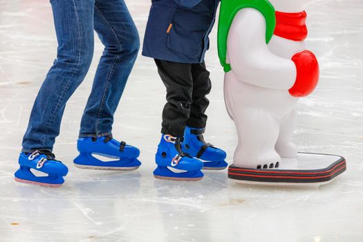 Winter day at the skating rink. A small child learns to skate with the support of an adult parent and holding on to a toy helper. Sports lifestyle and leisure. Copy space, selective focus, close-up.