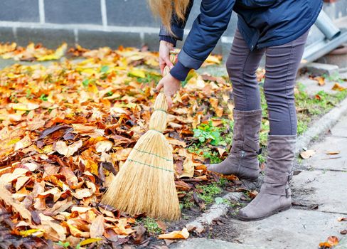 The hostess sweeps away fallen leaves from the path with a broom in the autumn garden, selective focus.