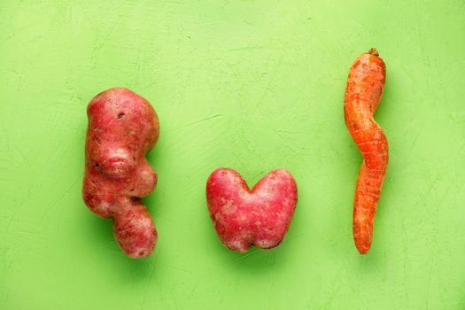 Ugly funny vegetables, lumpy potatoes in the shape of a heart and letters and a twisted carrot on a green plaster background. Coarse vegetables or food waste concept. Top view, copy space.