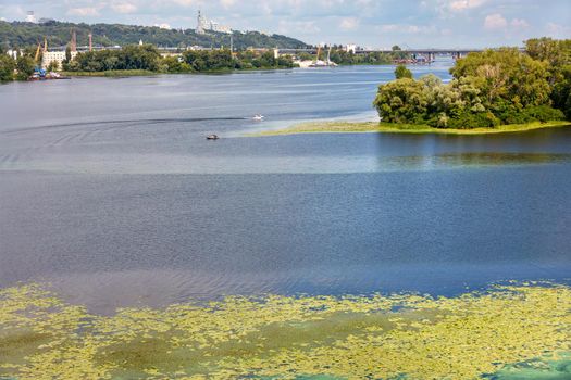 The most beautiful river Dnipro with river lilies floating on the water and fishing motor boats. In the background, one can see the slopes of the right bank of Kyiv with temples, monuments and bridges.