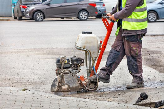 A road worker in a light green reflective vest smoothly cuts old asphalt with a diamond cut-off wheel on the road in front of a city street in a blurred form. Copy space.