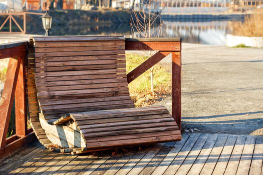 A wooden armchair with horizontal slats sits in a seating area by the river and is illuminated by the rays of the bright autumn sun.