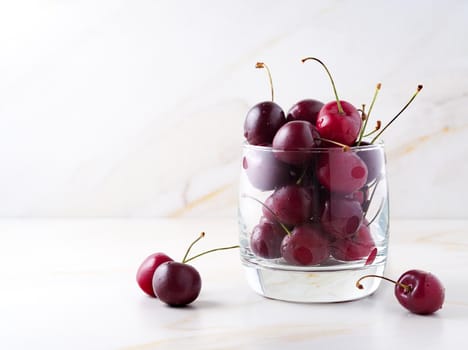 The red dark sweet cherries in glass on stone white table, side view, copy space