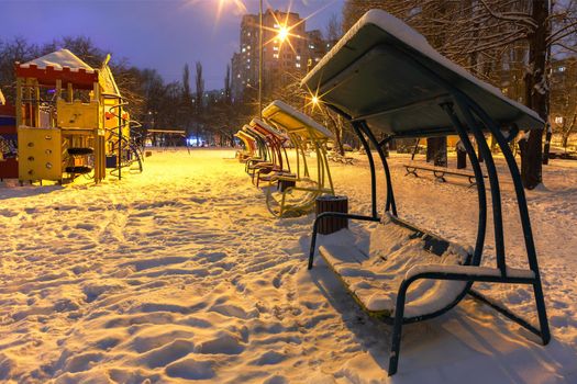 Empty playground with swinging wooden benches with metal railing in the snow against the blue twilight background on a winter evening in a city park and illuminated by the warm light of street lamps.