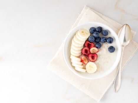 Large bowl of tasty and healthy oatmeal with fruits and berry for Breakfast, morning meal. Top view, white marble table, copy space