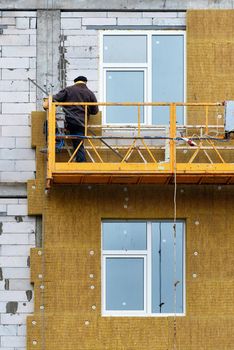 The builder insulates the facade of a house under construction with mineral insulation around the windows while standing on a construction suspended platform, which is a concept of saving heat in the house. Vertical image.