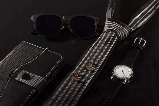 Notebook in leather cover, sunglasses, tie, cuff-links, watch with a leather strap on a black background