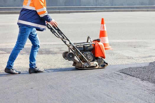 A road worker compacts asphalt on the carriageway with a petrol vibratory plate compactor.