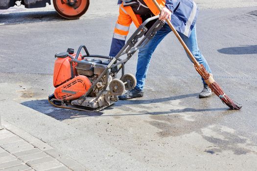 A road worker adjusts the operation of a petrol vibratory compactor against the background of a vibratory roller, and holds an old broom for asphalting a small section of the road. Copy space.