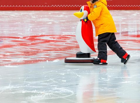 A child in a bright yellow jacket learns to skate on an ice rink holding on to an assistant, a toy penguin dressed in a red frock coat. Copy space, selective focus, close-up.