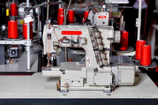 Professional multifunctional and modern sewing machine with spools of red thread close-up on the background of a sewing workshop in blur.