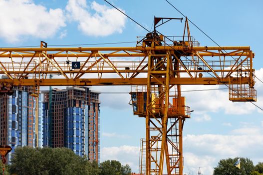 Metal construction crane for moving heavy loads against the background of residential buildings under construction in blur and blue sky. Copy space.