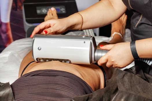 A professional cosmetologist performs an electromagnetic massage of the abdominal muscles on a woman patient on a high-intensity simulator.