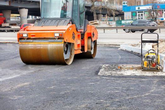 A heavy road vibratory roller and a petrol vibratory compactor plate are used to compact fresh asphalt on a new stretch of road. Copy space.