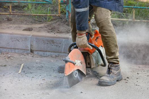 A worker uses a portable petrol saw and a diamond cutting blade to cut old asphalt in a cloud of dust, sand and flying sparks. Close-up, copy space.