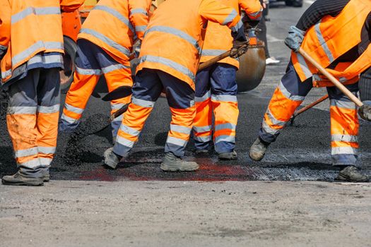 A team of road workers in orange reflective uniforms refurbish a section of the road with fresh asphalt and level it with shovels before compacting with a road roller. Clear day, copy space.