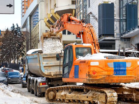 A large metal bucket of an orange tracked excavator loads snow from a city road onto a truck body against a backdrop of a city street and passing cars.