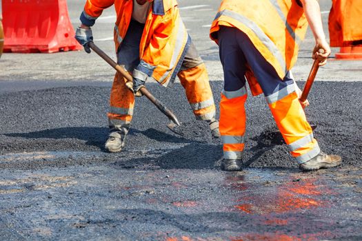 A team of road workers in orange overalls use shovels to level the fresh asphalt to evenly distribute the road surface on the roadway. Copy space.