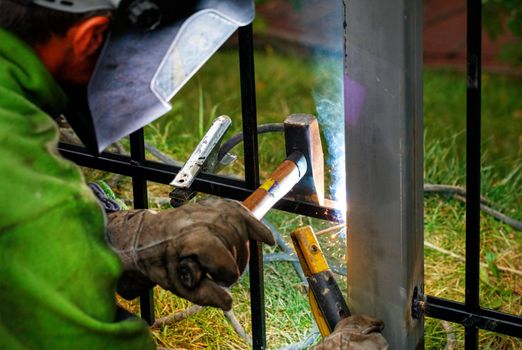 A welder wearing a safety helmet and gloves is welding a metal fence, bright sparks fly, blue smoke, selective focus, copy space.